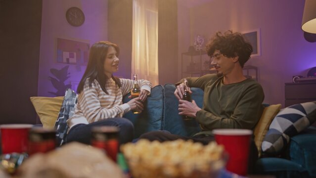Young boy and girl are having pleasant talk on sofa. Two friends are looking eye to eye and drinking bottles of beverage. Two young people are share thoughts at background of well-designed room.