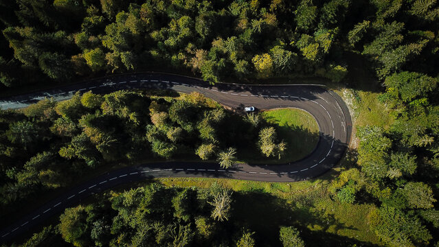 Aerial view of a winding mountain road cutting through a dense forest with a single car navigating a sharp curve in Mijoux, Auvergne-Rhone-Alpes, France.