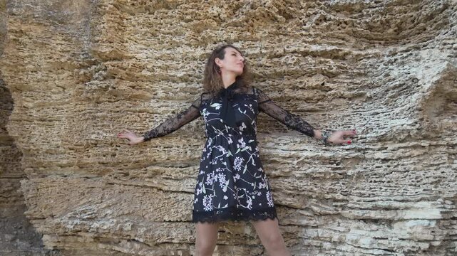 Woman nature pose against rough rock wall wearing a floral dress, outdoors in sunlight expressing freedom.