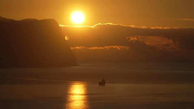 Sunrise ocean ship Silhouette of a fishing vessel at sea under a vibrant golden sky with majestic coastal cliffs
