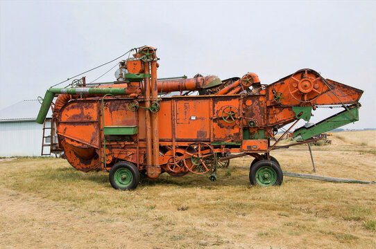 A side view of a vintage mechanical threshing separator with rusty orange paint and green wheels. The historic agricultural machine stands in a farmyard next to a white metal shed.