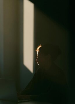 Silhouette of a person sitting in front of a laptop computer. the person is facing away from the camera, with their head turned towards the right side of the image.