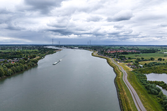 Aerial view of the Lek river flowing through the landscape with a cargo barge, green banks, a road, and power lines under a cloudy sky in Kinderdijk, Zuid-Holland, Netherlands.