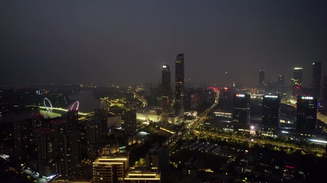 Aerial night view of a modern urban area featuring tall skyscrapers, illuminated roads, and a bridge over a river.