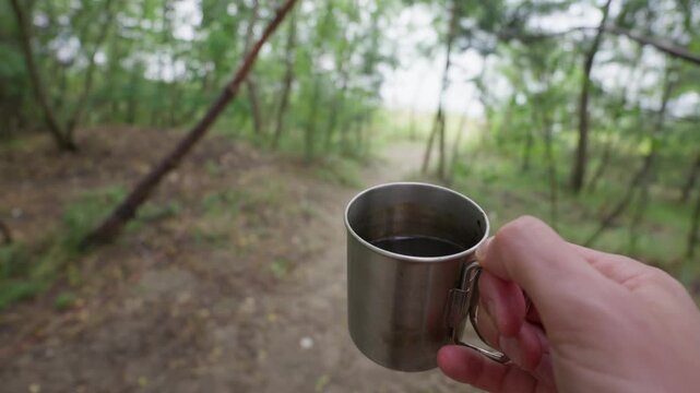 A person holds a stainless steel camping mug filled with black coffee while standing on a dirt forest trail. The traveler pauses during a hiking journey to enjoy a warm drink in nature.