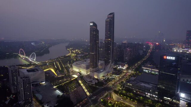A twilight aerial view of a modern urban area featuring twin skyscrapers, a waterfront, and illuminated city infrastructure.