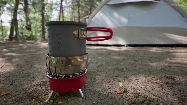 A portable camping stove burns brightly under a metal pot filled with water on the sandy ground. A large dome camping tent stands in the background surrounded by green trees.