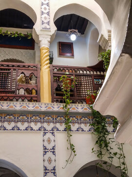 Traditional Moroccan riad interior featuring ornate wooden railings vibrant ceramic tiles intricate plasterwork arches and cascading green ivy plants illuminated by soft warm hanging lights