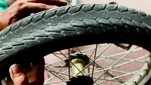 A close-up of a person's hand holding a heavily damaged bicycle tire with a large tear or hole in the sidewall. Showing a flat tire needing repair or replacement on a concrete ground.