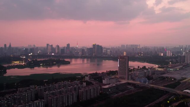 A panoramic urban view at dusk, featuring a calm lake reflecting the pink-hued sky and surrounding city buildings.