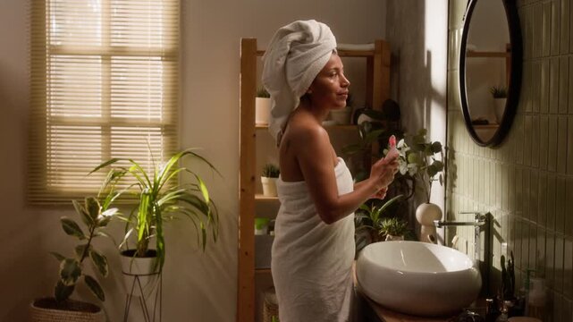 Hispanic young woman wrapped in towel using pink LED light therapy facial massager in bathroom. Footage is perfect for beauty technology commercials and skincare brand marketing