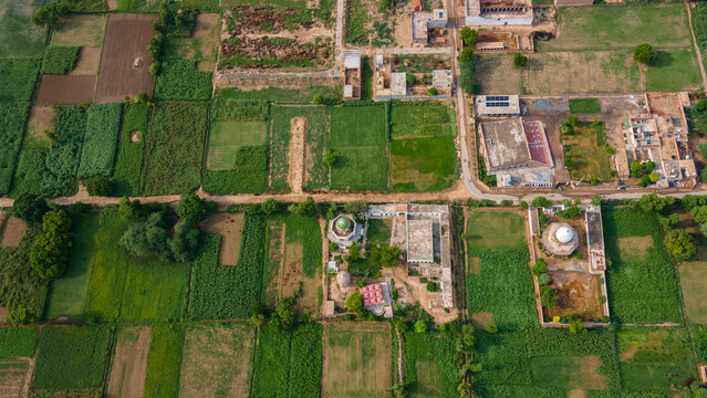 Aerial view of a rural village with lush green agricultural fields, traditional houses with flat roofs, small shrines with domes, and dirt paths connecting the properties in Punjab, Pakistan.