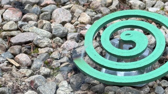 Turquoise mosquito repellent coil lies on smooth gray stones in an outdoor setting, viewed from above with its spiral shape clearly visible, pest control advertising, outdoor living, camping products,