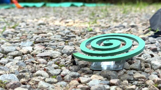 Turquoise mosquito repellent coil sits low on rounded gray pebbles outdoors, showing its spiral form against a natural ground surface, pest control advertising, outdoor living, camping products, insec