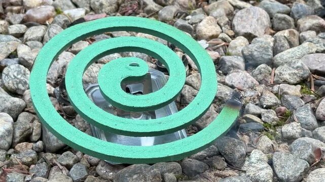 Turquoise spiral mosquito repellent coil rests on rounded gray pebbles outdoors, pest control advertising, outdoor living, camping products, natural protection, summer outdoors, insect prevention