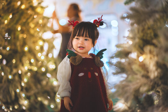 Happy toddler girl wearing reindeer costume among illuminated Christmas trees, festive holiday celebration