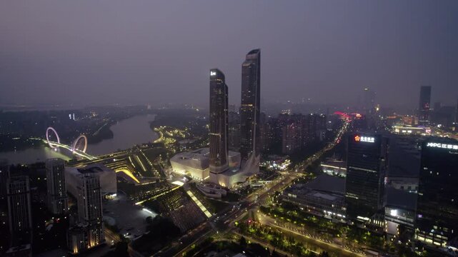 A twilight aerial view of a modern urban district with twin skyscrapers by a river, illuminated at night.
