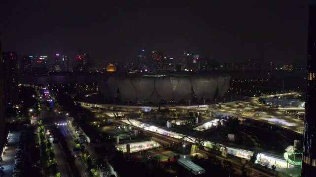 A night aerial view of a large stadium complex with illuminated pathways and distant urban skyline in the background.