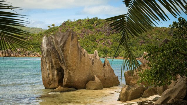 Scenic tropical coastline in the Seychelles with dramatic granite rocks, turquoise sea, lush green hills and palm leaves.