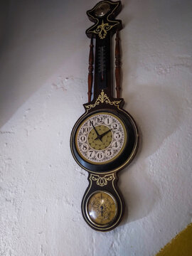 Vintage wooden wall clock featuring an integrated thermometer and barometer with decorative gold accents hanging on a white textured plaster surface in soft natural interior lighting