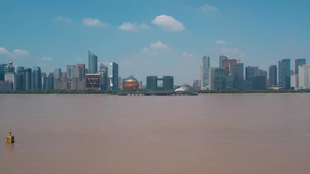 A wide muddy river stretches toward a modern urban skyline with distinctive buildings under a bright blue sky with clouds.