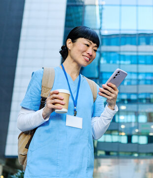 Nurse in scrubs enjoying coffee and using a mobile phone outside a modern medical facility during a sunny day