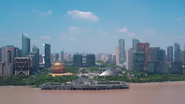 A panoramic daytime view of a modern urban skyline with skyscrapers lining a muddy river, green spaces and distinctive buildings under a clear blue sky.