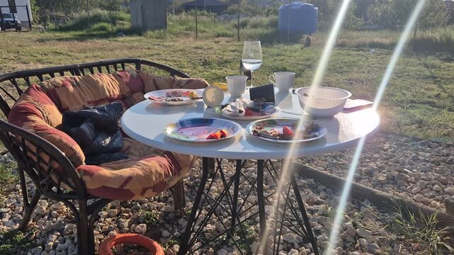 Outdoor dining table remains set with leftover breakfast snacks in a garden backyard during bright golden hour. Static wide shot.