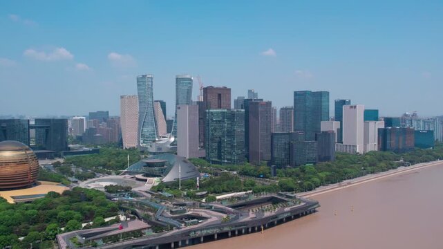 Aerial view of modern urban skyline with skyscrapers along a brown river, surrounded by green parks under clear blue sky.