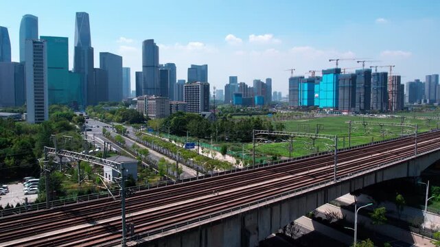 A high speed train travels on an elevated railway viaduct in a modern urban area with skyscrapers and construction sites nearby.
