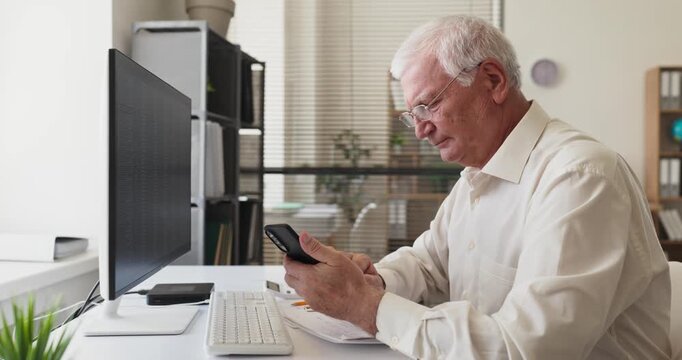 Senior accountant man using smartphone in office. At the desk with computer and documents, he reviews figures and performs data analysis. Clear finance concept for business communication needs.