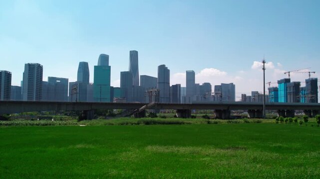 A modern urban skyline with high-rise buildings, an elevated concrete bridge, and a green field under a clear blue sky in daytime.
