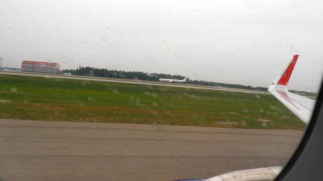 Commercial airplane view through rainy window looking at distant plane accelerating on runway during overcast day at airport, wing visible in frame.