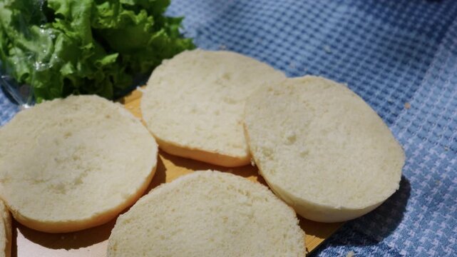 Sliced burger buns and fresh green lettuce rest on a wooden board placed over a blue patterned picnic blanket outdoors. Concept of outdoor dining, food preparation, and summer meal making.