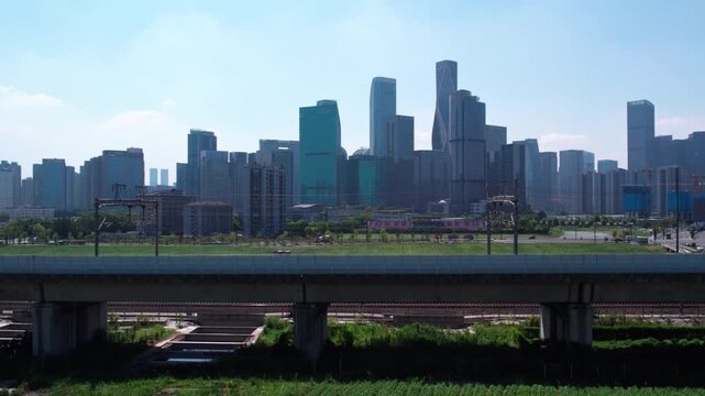 A daytime city view featuring a modern skyscraper skyline, an elevated overpass, and a green open space in the foreground.