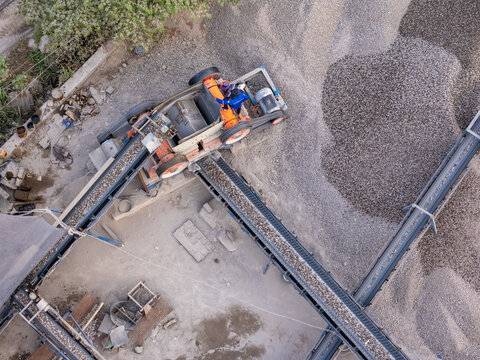 Aerial view of a stone crushing machine with a conveyor belt processing gravel piles and a worker standing on the machinery in Klaten, Central Java, Indonesia.