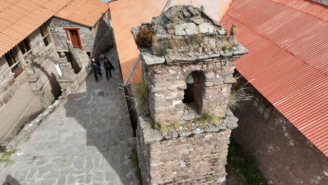 Aerial view of the stone bell tower on Taquile Island with a cobblestone path and people walking past traditional buildings with orange roofs in Huillanopampa, Puno, Peru.