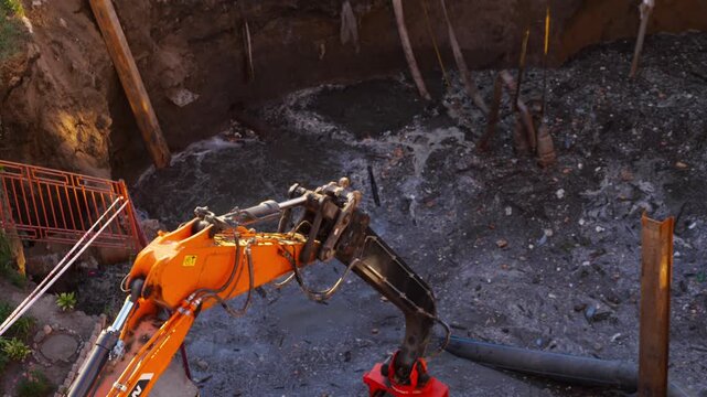 Excavator bucket working inside deep excavation pit repairing sewer collector pipe failure with dirty wastewater flow, high angle shot.