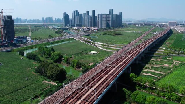 An elevated high-speed railway with a train running through, adjacent to urban buildings and green fields.