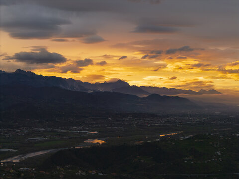 Aerial view of the Apuan Alps at sunset with a winding river flowing through the valley under a dramatic golden sky in Tuscany, Italy.