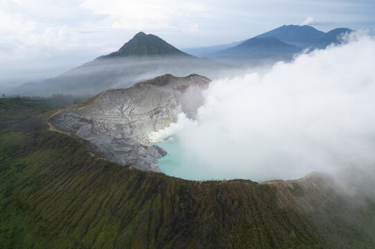 Aerial view of Ijen volcano crater with its turquoise acidic lake and rising sulfur smoke surrounded by lush green mountain slopes in East Java, Indonesia.