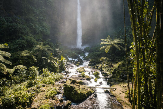 Aerial view of Kapas Biru Waterfall cascading into a rocky stream surrounded by lush tropical vegetation and towering cliffs in the dense forest, Kecamatan Pronojiwo, Jawa Timur, Indonesia.
