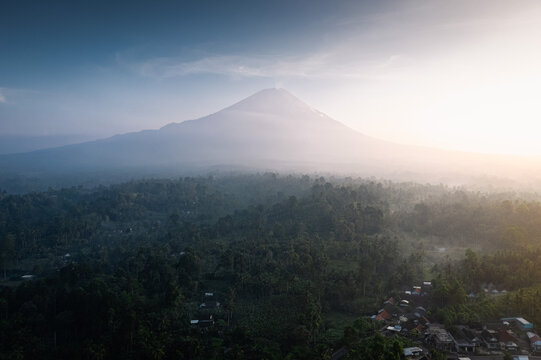 Aerial view of Mount Semeru rising above the lush tropical forests and small village in the morning light in Lumajang, East Java, Indonesia.