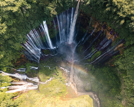 Aerial view of Tumpak Sewu Waterfall featuring cascading streams, lush tropical vegetation, and a vibrant rainbow in the mist in Kecamatan Pronojiwo, Jawa Timur, Indonesia.