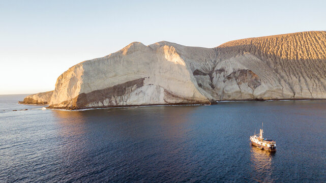 Aerial view of San Benedicto Island with its dramatic volcanic cliffs rising from the deep blue ocean and a small boat anchored nearby during the golden hour in Colima, Mexico.