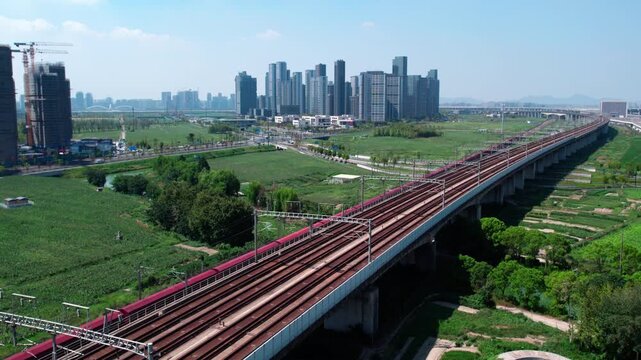 An elevated railway bridge runs through a developing urban area with green spaces and high-rise buildings under clear skies.