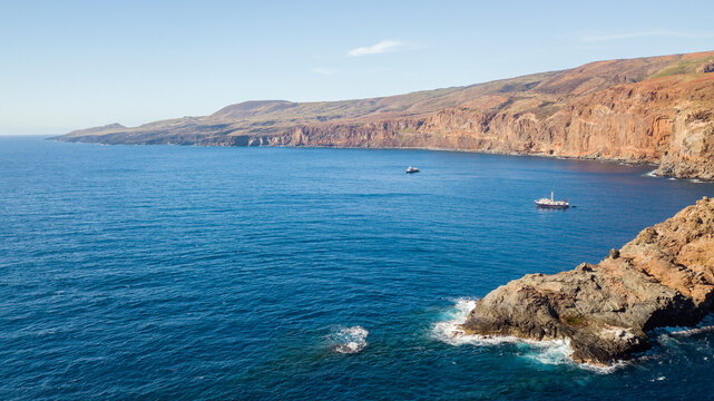 Aerial view of Socorro Island with rugged volcanic cliffs, deep blue ocean waters, and two boats anchored near the rocky coastline in Colima, Mexico.