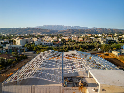 Aerial view of a large steel frame roof structure under construction at a building site, surrounded by urban residential areas, olive groves, and distant mountains in Chania, Greece.