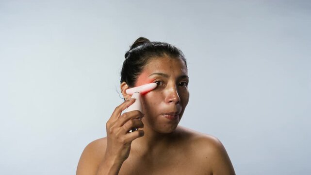 Portrait of young Hispanic woman using pink LED light therapy facial massager on her skin. Clip is perfect for beauty e-commerce ads and skincare product marketing campaigns