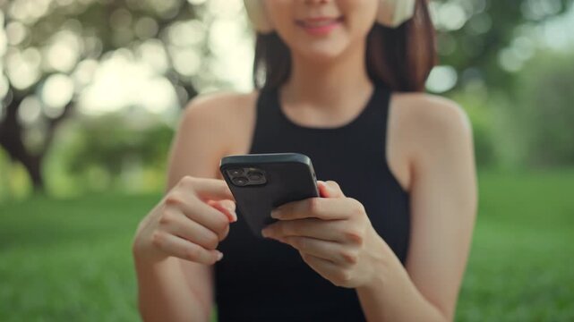 Young Asian woman using smartphone while wearing wireless headphones outdoors, digital lifestyle and mobile entertainment in park